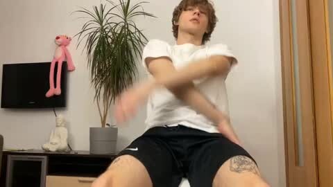 Media: Video of a young man with curly hair, wearing a white t-shirt and black shorts, sitting with crossed legs, in a modern living room with a pink stuffed toy, potted plant, and wooden door.