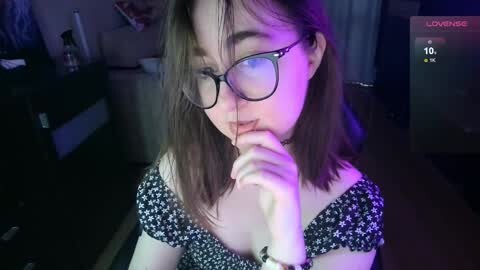 Media: Video of a young woman with shoulder-length brown hair, wearing glasses, a black floral dress, and biting her finger in a dimly lit room, with a \"Lovingly\" watermark in the top right corner.