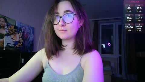 Media: Video of a young woman with shoulder-length brown hair, glasses, and a grey tank top, smiling, in a dimly lit room with colorful posters and a digital clock displaying 20:50.