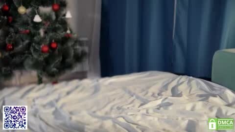 Media: A video of a neatly made bed with white sheets and a dark blue curtain backdrop. A Christmas tree with red and gold ornaments is partially visible in the left background.