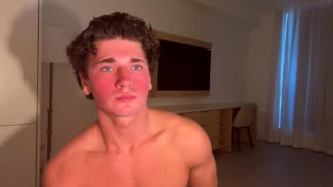 Media: Video of a shirtless young man with curly brown hair, red face, and blue eyes, standing in a modern, dimly lit room with beige walls, a flat-screen TV, and a window with sheer white curtains.