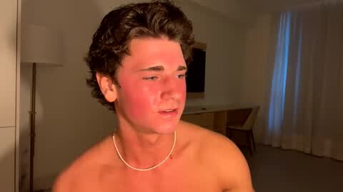 Media: Video of a young, shirtless man with curly brown hair and a red face, wearing a thin silver necklace. He stands indoors, next to a tall lamp and a wooden table with chairs.