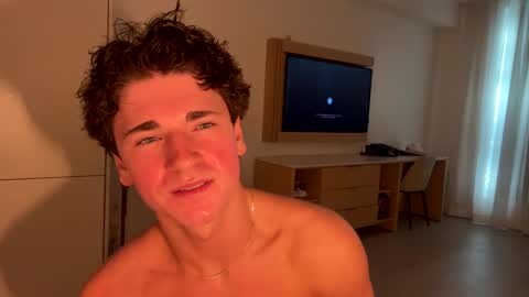 Media: Video of a shirtless young man with curly hair, smiling, standing in a dimly lit bedroom with beige walls, wooden dresser, and a TV displaying a blue screen.