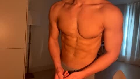 Media: Video of a muscular, shirtless male with fair skin, standing in a dimly lit room. He wears only briefs, highlighting his defined abs and pecs. Background shows beige walls and a partially visible window with sheer white curtains.
