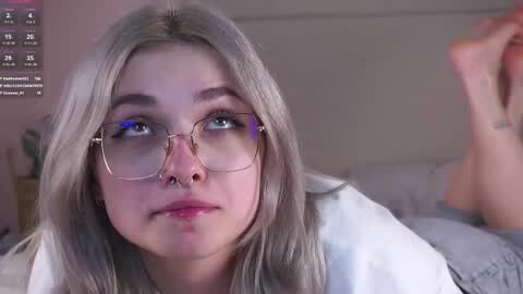 Media: Video of a young woman with fair skin, light gray hair, and blue eyes wearing glasses and a septum piercing. She is lying on a bed, looking contemplative.