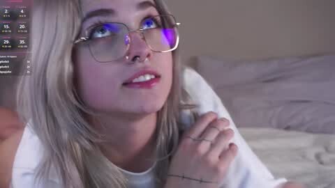 Media: Video of a young woman with light skin, blue eyes, and long blonde hair, wearing glasses, a septum piercing, and a white shirt, lying on a bed with gray sheets.