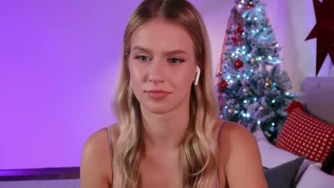 Media: Video of a blonde woman with light skin and wavy hair, wearing a white tank top, sitting in front of a decorated Christmas tree with purple lighting and red ornaments.