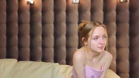 Media: Video of a young Caucasian woman with light skin and blonde hair in a bun, wearing a pink camisole, sitting on a beige bed with a tufted brown headboard.