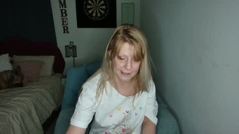 Media: Video of a smiling, blonde-haired woman in a white floral shirt sitting on a teal couch in a dimly lit room with a darts board and a bed in the background.