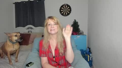 Media: A blonde woman in a red plaid shirt waves in a cozy bedroom with a beige dog, a dartboard, and a Christmas tree.