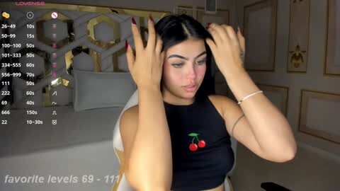 Media: Video of a young woman with long black hair, wearing a black crop top with cherry print, adjusting her hair in a modern, luxurious bedroom.