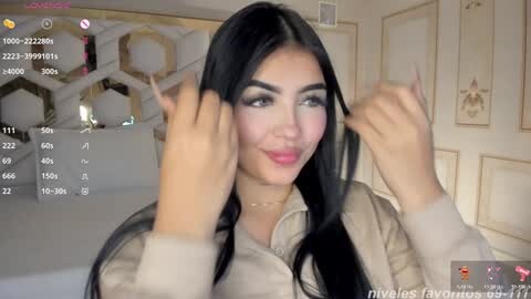 Media: A video of a young woman with long black hair, wearing makeup and a beige jacket, playfully touching her hair in a bedroom with a white bed and gold-patterned wall.