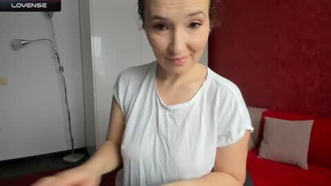 Media: Video of a smiling, fair-skinned woman with brown hair tied back, wearing a white T-shirt, standing in a modern room with red walls, a red sofa, and a floor lamp.