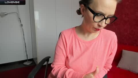 Media: Video of a slim, light-skinned woman with short brown hair and black glasses, wearing a pink long-sleeved shirt, sitting on a red chair in a modern room with white walls and red wallpaper.