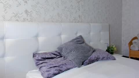 Media: Video of a minimalist bedroom with a white tufted headboard, light purple bedding, and a wicker chair in the corner. The wallpaper features a subtle geometric pattern.