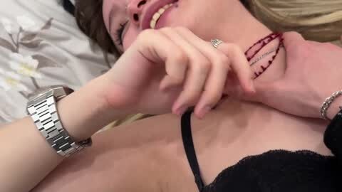 Media: Video of a smiling, fair-skinned woman with long blonde hair, wearing a black lace bra, a red beaded necklace, and a silver wristwatch, lying on a floral-patterned bed.
