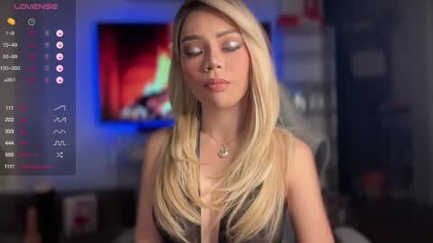 Media: Video of a blonde woman with long, straight hair, wearing a low-cut black dress, standing in a dimly lit room with a TV displaying a red and blue screen.