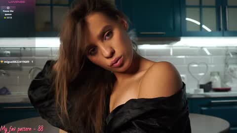 Media: Video of a young woman with light brown skin and long brown hair, wearing a black off-shoulder top, in a modern kitchen with teal cabinets and a white backsplash.