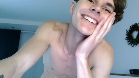 Media: Video of a smiling, shirtless, young white male with fair skin and short brown hair, resting his chin on his hand in a casual indoor setting with a Christmas wreath in the background.