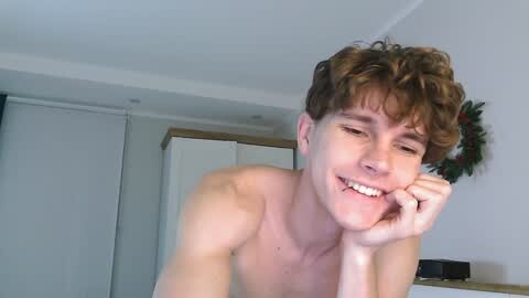 Media: Video of a young, shirtless man with curly brown hair, smiling, leaning on his elbow, in a modern, minimally decorated room with white walls and a Christmas wreath.