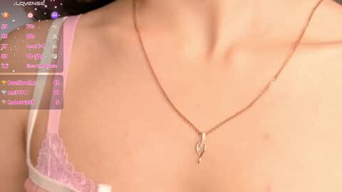 Video of a woman's upper torso in a pink lace bra, wearing a thin gold necklace, with chatroom text in the background.