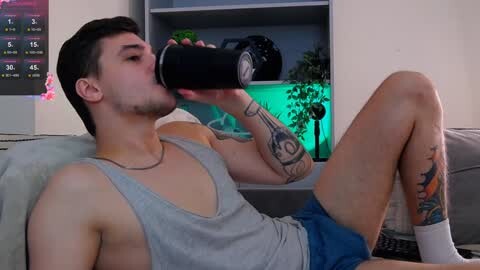 Media: Video of a fit, tattooed man in a grey tank top and blue shorts, drinking from a black water bottle, lounging on a couch, with a digital TV displaying sports scores in the background.