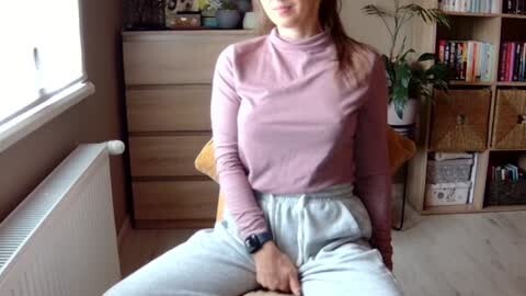 Media: Video of a young woman with fair skin, brown hair, wearing a pink turtleneck and gray sweatpants, seated on a wooden chair in a cozy, bookshelf-filled room with a window and potted plant.