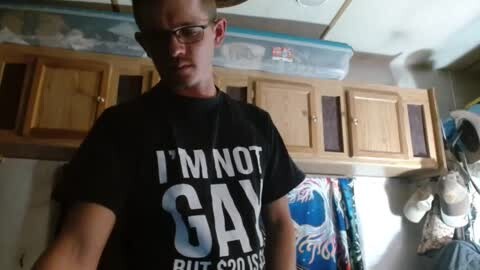 Media: Video of a young man with glasses, short brown hair, wearing a black T-shirt with \"I'M NOT GAY BUT GOD IS\" text, in a RV kitchen with wooden cabinets and a colorful cloth hanging.