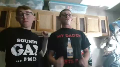 Media: Video of two young, fair-skinned men with glasses, one with red hair, the other with short hair, wearing black T-shirts with \"SOUNDS GAY?... I'M GAY!\" and \"MY DADDY,\" respectively, standing in a cluttered, dimly lit storage room with wooden cabinets and plastic bins.