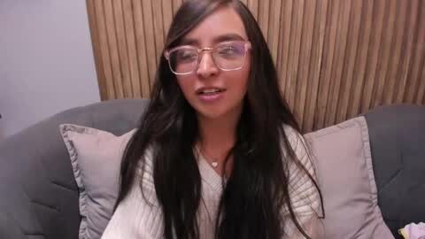 Media: A video of a young woman with long, straight black hair, wearing glasses and a beige sweater, seated on a grey couch with beige pillows, against a wooden background.