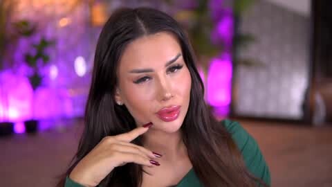 Media: Video of a light-skinned woman with long, straight dark hair, wearing a green top and red lipstick. She has well-defined eyebrows, manicured nails, and a confident expression. Background features blurred greenery and purple lighting.