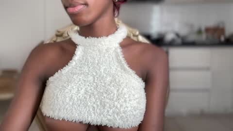 Media: Video of a smiling, dark-skinned woman with braided hair, wearing a white, textured, halterneck crop top. Background shows a modern kitchen with white cabinets and countertops.