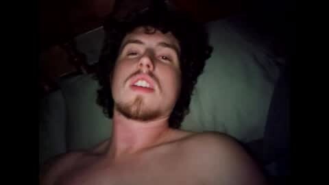 Media: Video of a shirtless, light-skinned man with curly black hair and a trimmed beard, lying on a bed with a green pillow, in a dimly lit room.