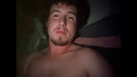 Media: Video of a smiling, topless white man with curly hair and a trimmed beard, lying on a bed with dark sheets and a pillow.