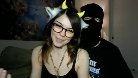 Media: Video of a young woman with glasses, wearing a black tank top and yellow devil horns, smiling. A man in a black mask and shirt is behind her. Background includes a wall with abstract art and a lamp.