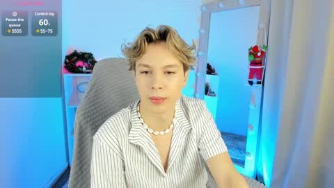 Media: Video of a young, fair-skinned man with wavy, shoulder-length blonde hair, wearing a striped shirt and pearl necklace, seated in a dimly lit room with a pink flower arrangement and a red dress hanging in the background.