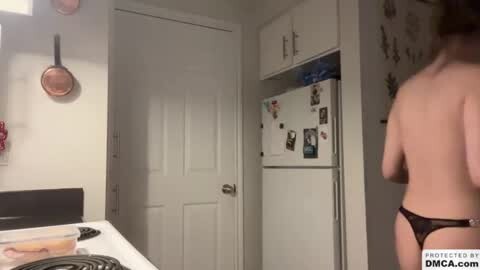 Media: A video of a naked woman wearing a black thong, standing in a kitchen with white cabinets, a refrigerator, and a stove.