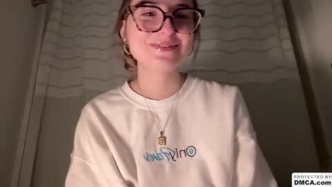 Media: Video of a young woman with glasses, light skin, brown hair, wearing a white hoodie with \"HOTWHEELS\" in blue and red, and a necklace, standing in a dimly lit bathroom with a wavy shower curtain.