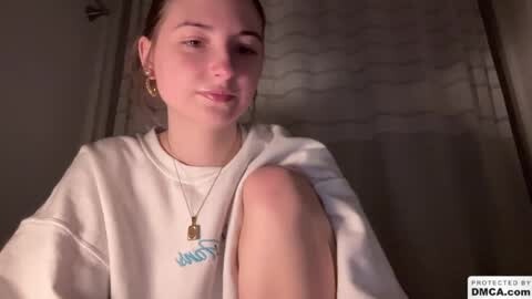 Media: A video of a young woman with fair skin and brown hair, wearing a white hoodie with blue text, sitting on a bed with a wavy curtain in the background.