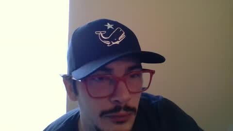 Media: Video of a man with medium skin tone, wearing a black baseball cap with a white whale logo, red glasses, and a dark shirt.