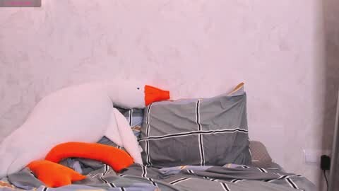 Media: Video of a white plush penguin with orange beak and feet, lying on a grey geometric-patterned bedspread with pillows, against a white textured wall.