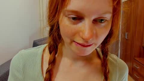 Media: Video of a young Caucasian woman with fair skin and red braided hair, wearing a light green ribbed sweater. She has a neutral expression, sitting in a room with wooden furniture and a white wall in the background.
