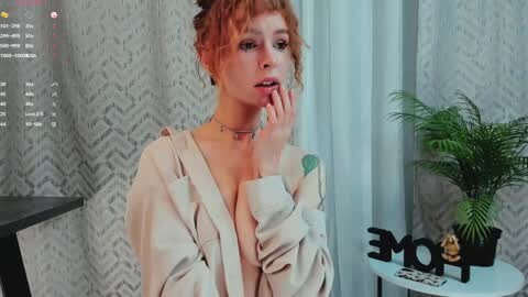 Media: A young woman with red hair in a messy bun, wearing a beige robe, stands in a modern room with gray patterned wallpaper, a potted plant, and a MOM sign on a white table.