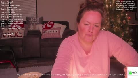 Media: Video of a middle-aged woman with fair skin and brown hair, wearing a pink shirt, sitting in a cozy living room with a Christmas tree and festive pillows in the background.