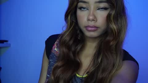 Media: Video of a young woman with wavy brown hair, wearing a black shirt, against a blue backdrop, looking pensive.