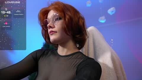 Media: A video of a fair-skinned woman with short, wavy red hair, wearing large, round glasses and a sheer black top, sitting in a plush chair in a dimly lit room with blue lighting and a digital clock displaying 10:45.