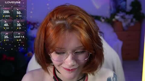 Media: Video of a fair-skinned woman with red hair, wearing glasses and a white top, smiling softly. Background shows a dimly lit room with a Christmas tree and a \"LoveLive!\" game overlay.