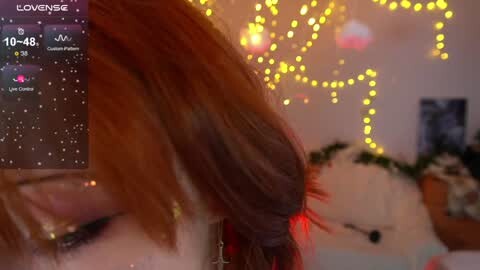 Media: A video of a woman with long, auburn hair, wearing a red bow, captured from behind in a cozy, dimly lit room adorned with string lights. The background features a plush toy and a framed picture.