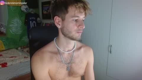 Video of a shirtless young man with light skin and short, tousled hair, wearing multiple pearl necklaces, sitting indoors near a white wardrobe. Background includes a partially visible green inflatable object and a black chair.