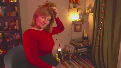 Media: Video of a smiling, red-haired woman with medium build in a red top, adjusting a floral headband in a cozy, dimly-lit room with bookshelves, candles, and a cluttered desk.